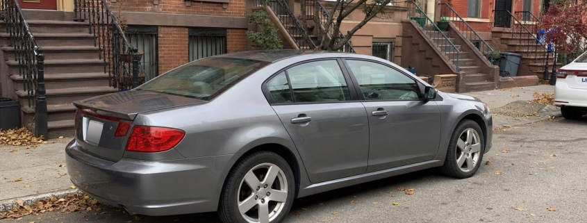 A car parked beside brownstone rowhouses on a Brooklyn residential street.