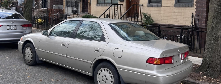 Silver sedan parked beside brownstone stoops on a Brooklyn residential street in autumn.