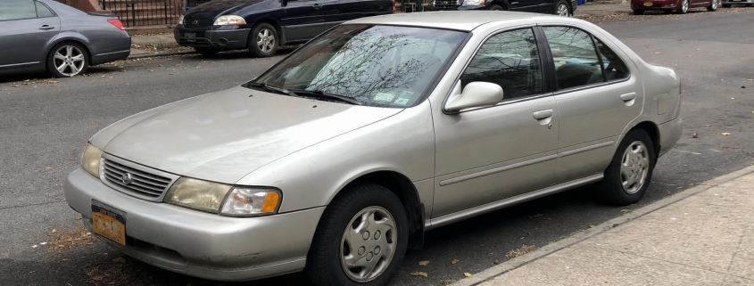 Silver sedan parked beside Brooklyn brownstones with stoops and autumn leaves along the curb.