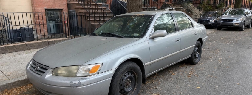 Silver sedan parked beside a Brooklyn sidewalk with brick rowhouses and brownstone stoops.