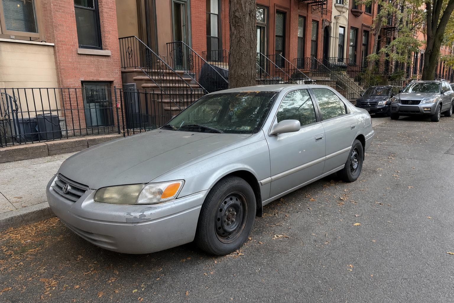 Silver sedan parked beside a Brooklyn sidewalk with brick rowhouses and brownstone stoops.