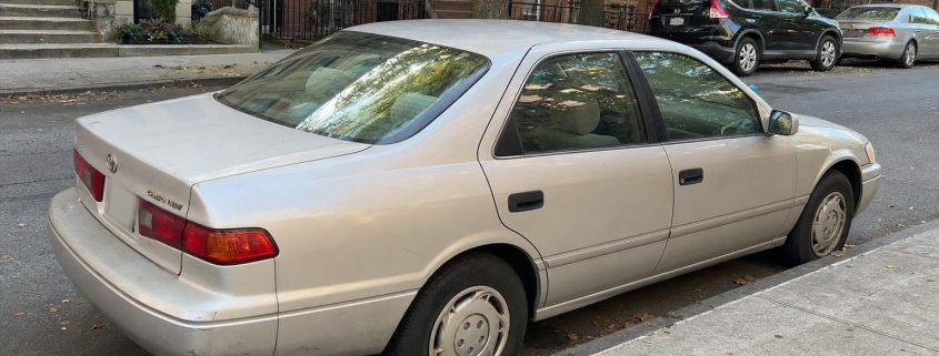 Silver sedan parked beside brownstone stoops on a Brooklyn residential street.