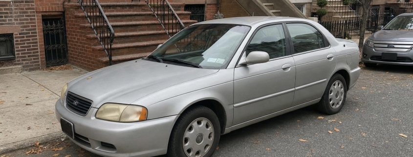 Silver sedan parked along a Brooklyn brownstone block with stoops and autumn leaves.
