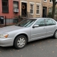 Older silver sedan parked beside Brooklyn brick rowhouses with stoops and autumn leaves along the curb.