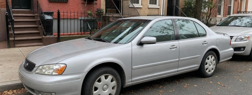 Older silver sedan parked beside Brooklyn brick rowhouses with stoops and autumn leaves along the curb.