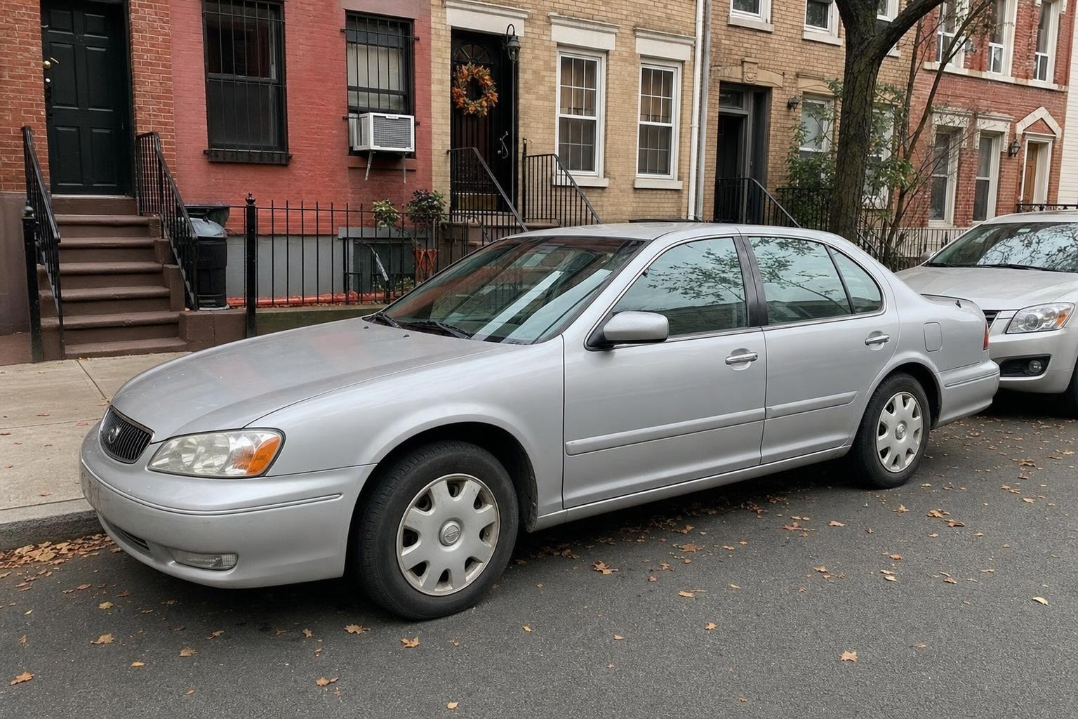 Older silver sedan parked beside Brooklyn brick rowhouses with stoops and autumn leaves along the curb.