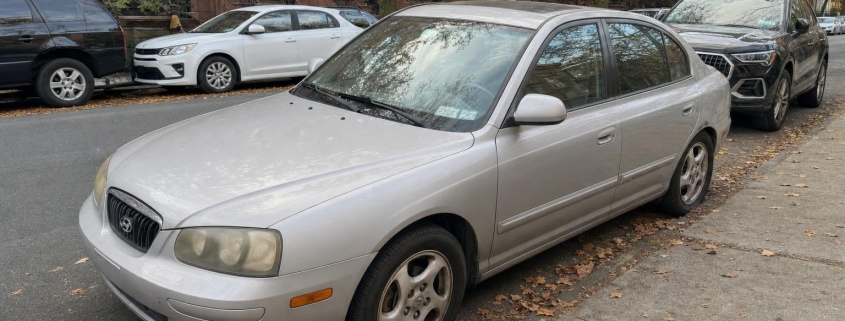 An older passenger car parked curbside beside Brooklyn brownstones with raised stoops.