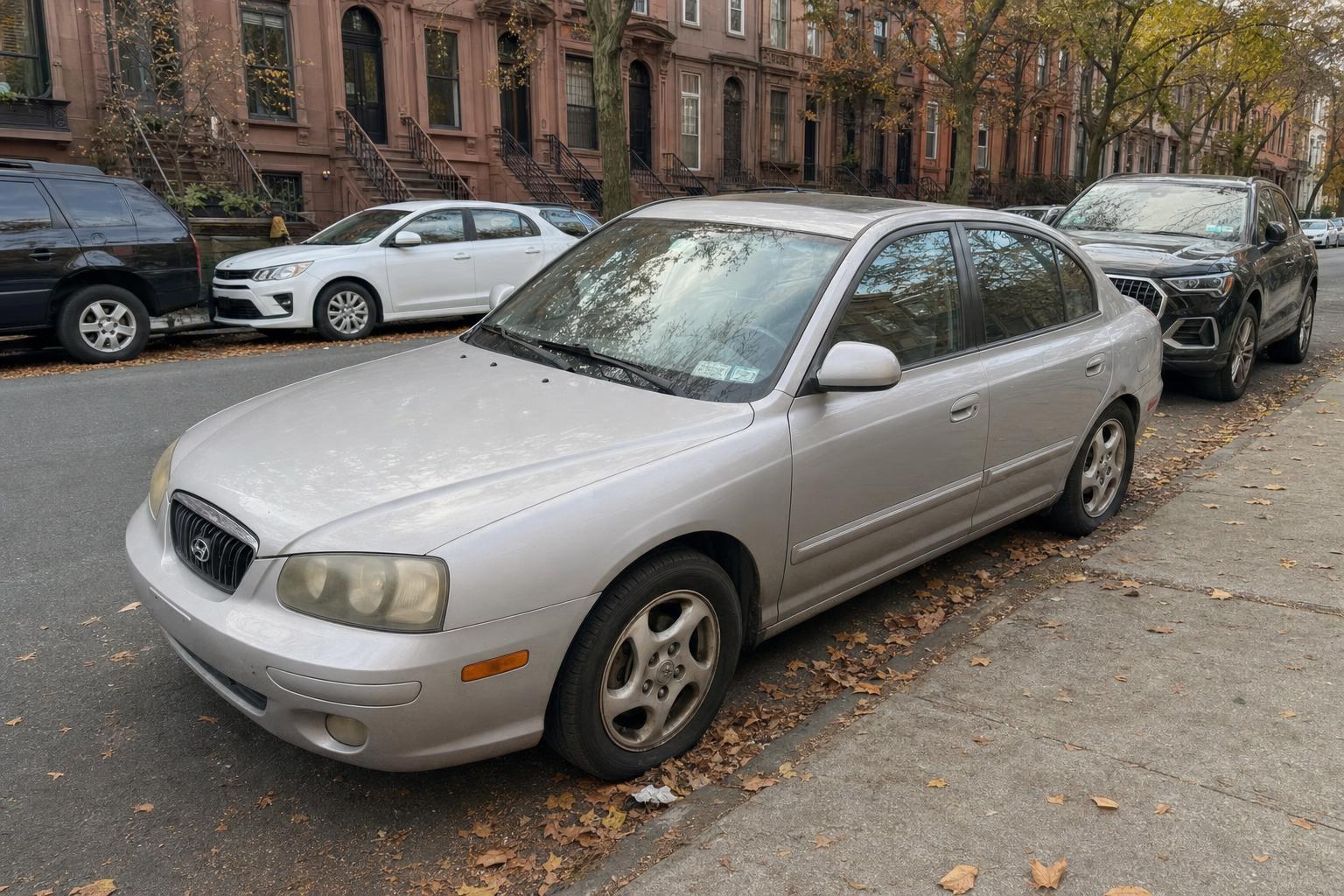 An older passenger car parked curbside beside Brooklyn brownstones with raised stoops.