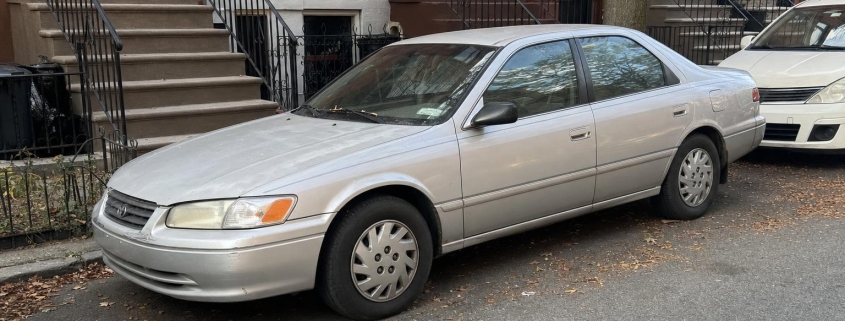 Silver sedan parked beside brick rowhouses and stoops on a Brooklyn residential street.