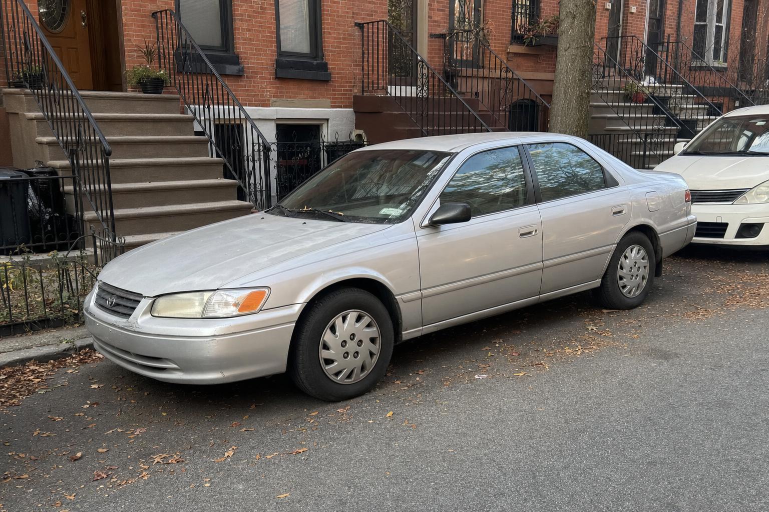 Silver sedan parked beside brick rowhouses and stoops on a Brooklyn residential street.