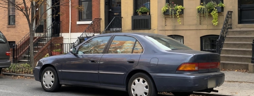 An ordinary used car parked along a Brooklyn brownstone block with stoops and curbside parking.