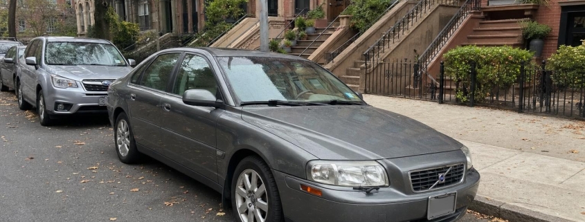 Used sedan parked beside brownstone stoops on a Brooklyn residential street in autumn.