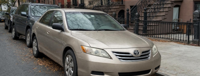 Older sedan parked beside brick rowhouses and brownstone stoops on a Brooklyn street in autumn.