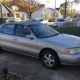 An older silver sedan parked in a residential driveway, with detached homes and mature trees in the background.