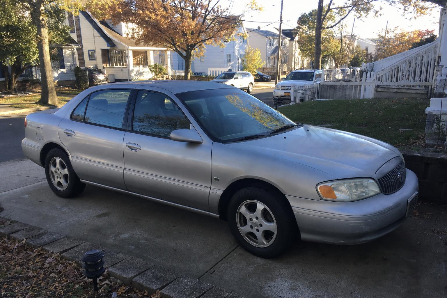 An older silver sedan parked in a residential driveway, with detached homes and mature trees in the background.