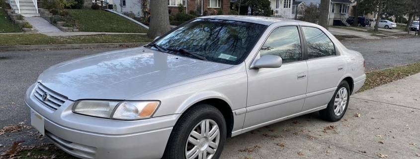 Older silver sedan in a Staten Island driveway beside detached homes and autumn trees.
