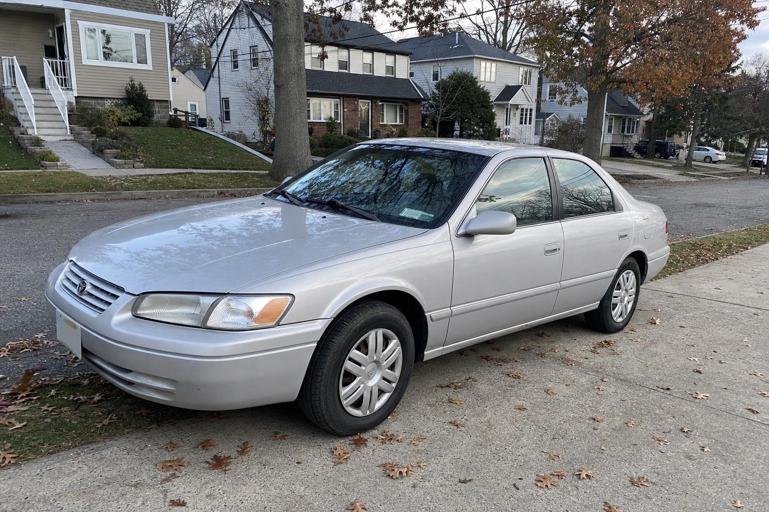 Older silver sedan in a Staten Island driveway beside detached homes and autumn trees.