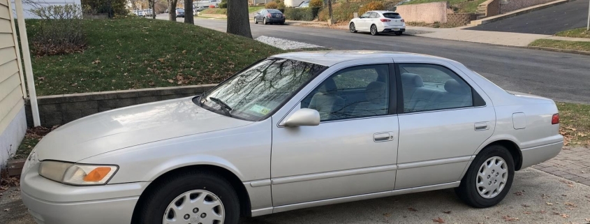 Older silver sedan in a Staten Island driveway beside detached homes and autumn trees