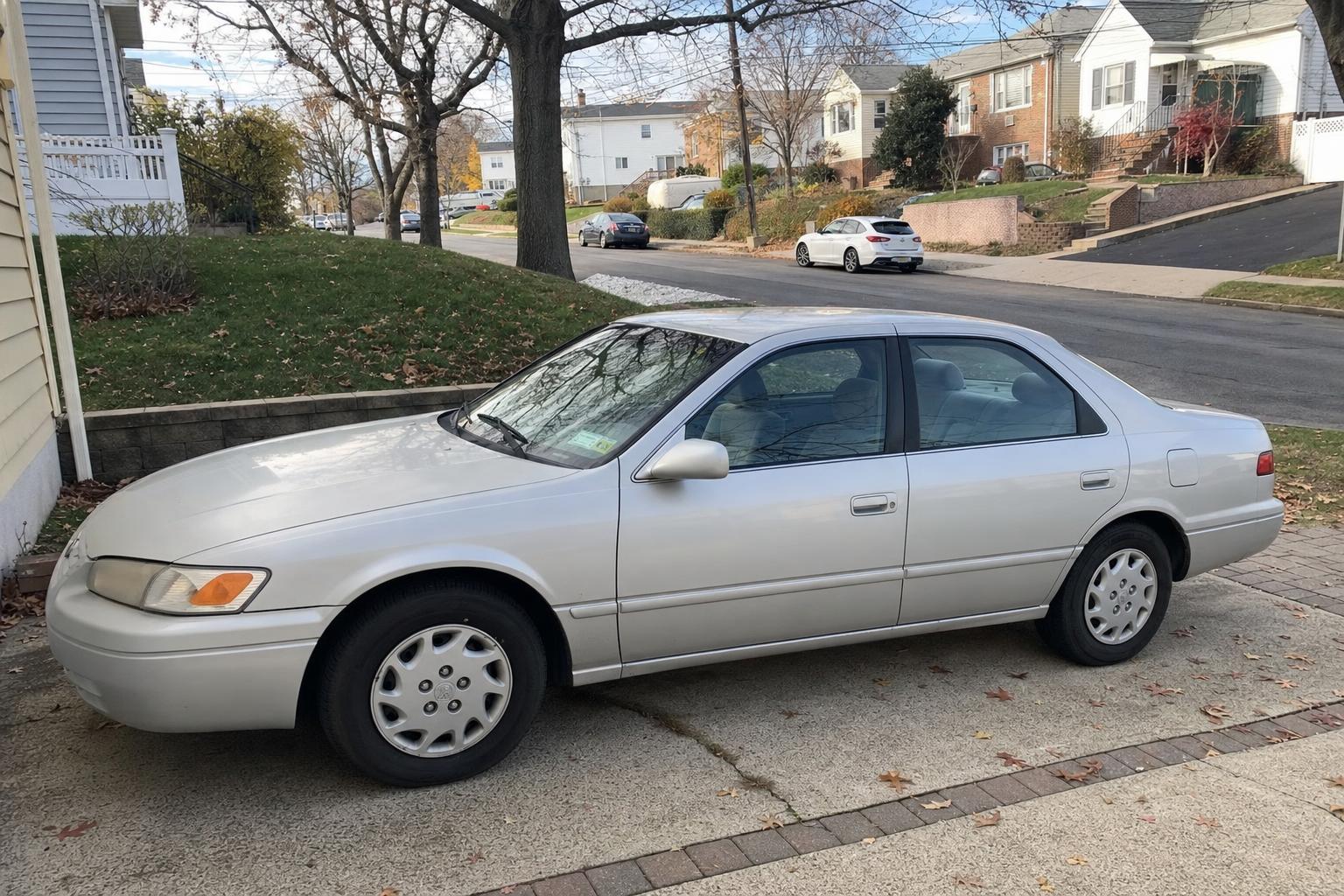Older silver sedan in a Staten Island driveway beside detached homes and autumn trees