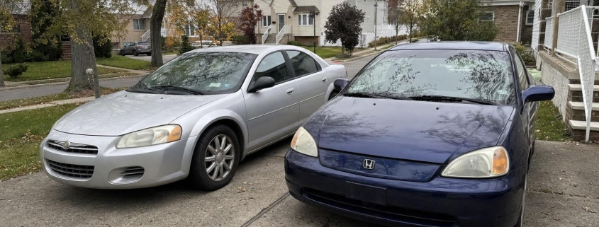 Two used cars parked side by side in a Staten Island residential driveway near detached homes.