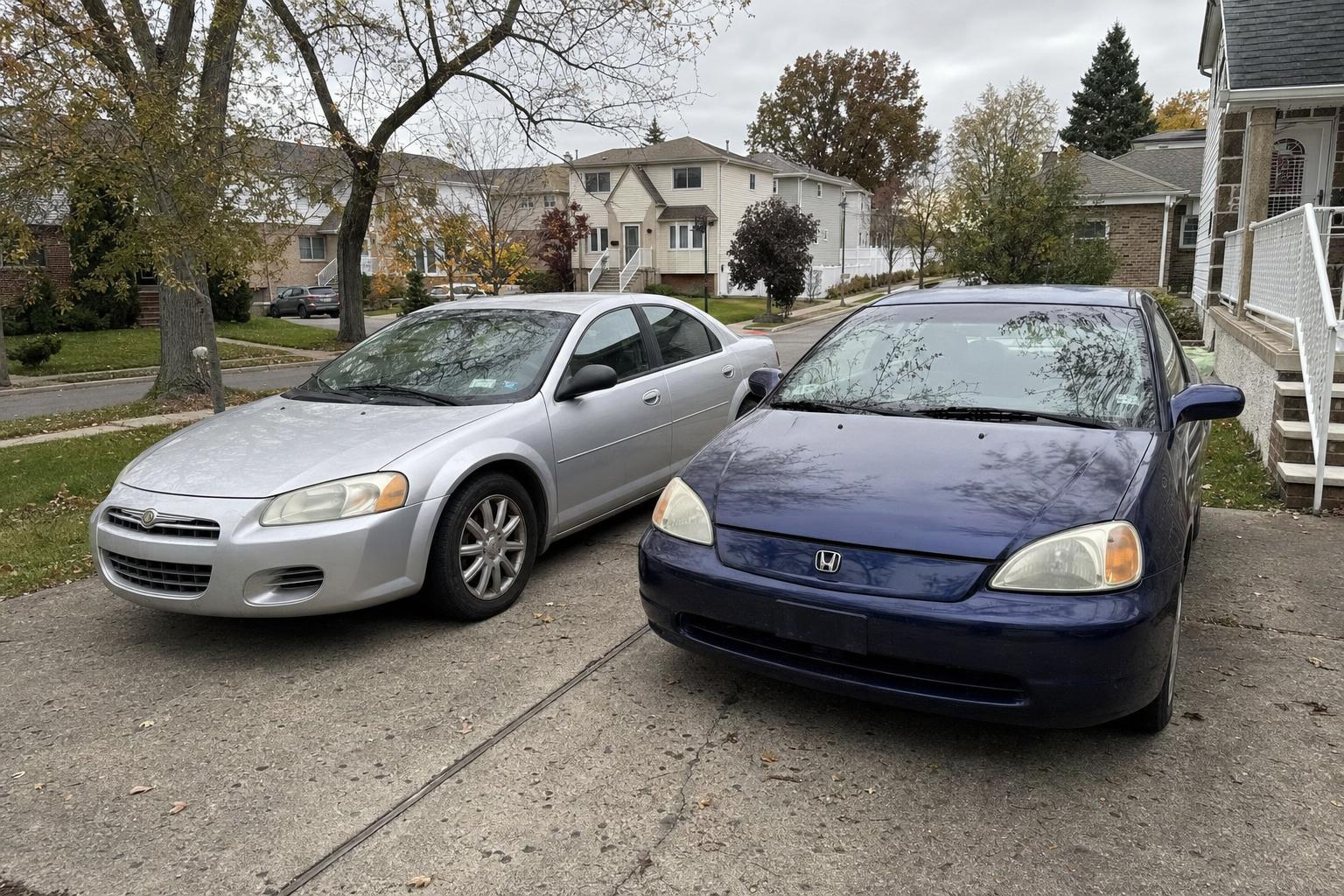 Two used cars parked side by side in a Staten Island residential driveway near detached homes.