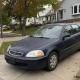 Older sedan parked in a Staten Island driveway beside detached homes and mature trees.
