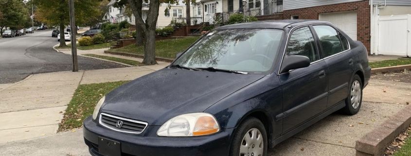 Older sedan parked in a Staten Island driveway beside detached homes and mature trees.