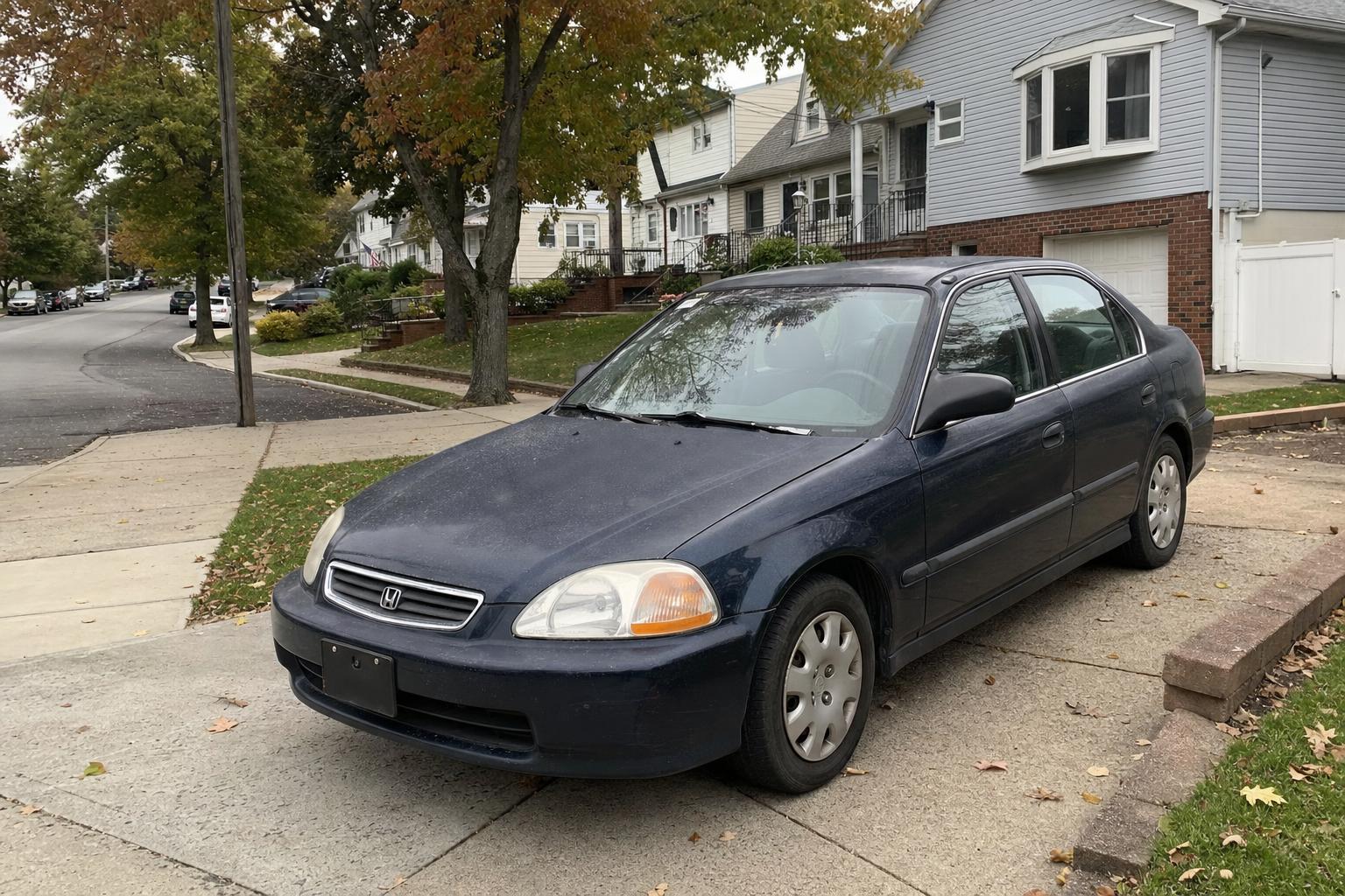 Older sedan parked in a Staten Island driveway beside detached homes and mature trees.