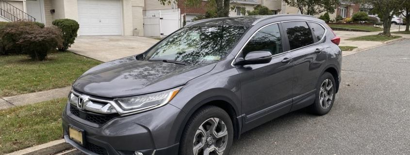 A gray Honda CR-V parked on a Staten Island residential street in front of detached homes and trees.