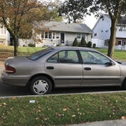 Shared Vehicle Donation Discussion in Staten Island An older sedan parked on a quiet Staten Island residential street lined with detached homes and trees.