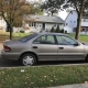 An older sedan parked on a quiet Staten Island residential street lined with detached homes and trees.