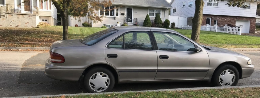 An older sedan parked on a quiet Staten Island residential street lined with detached homes and trees.
