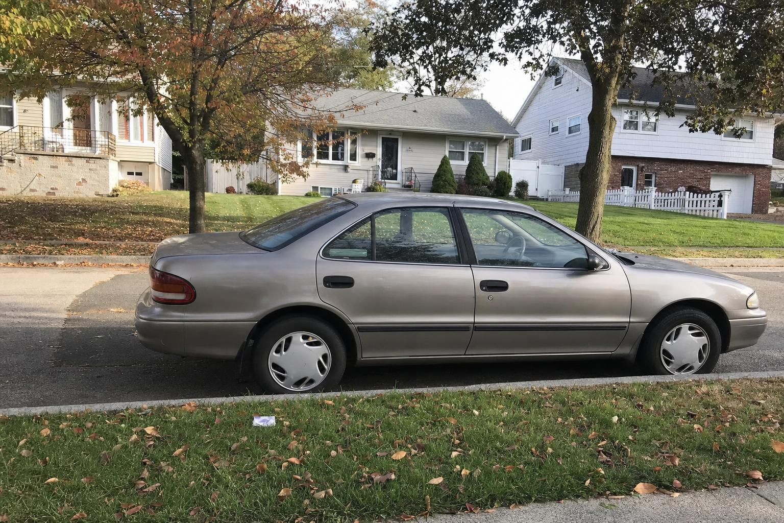 An older sedan parked on a quiet Staten Island residential street lined with detached homes and trees.